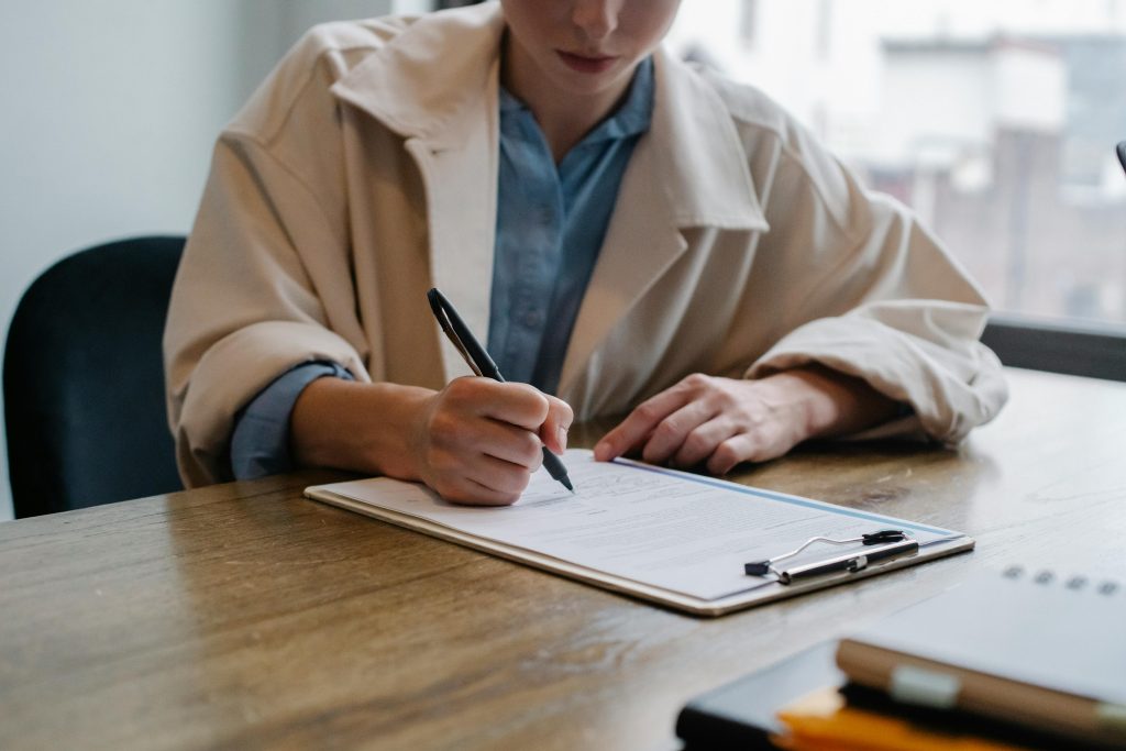 pexels-photo-5668869-5668869 A woman in a formal setting fills out paperwork on a clipboard at an office desk.